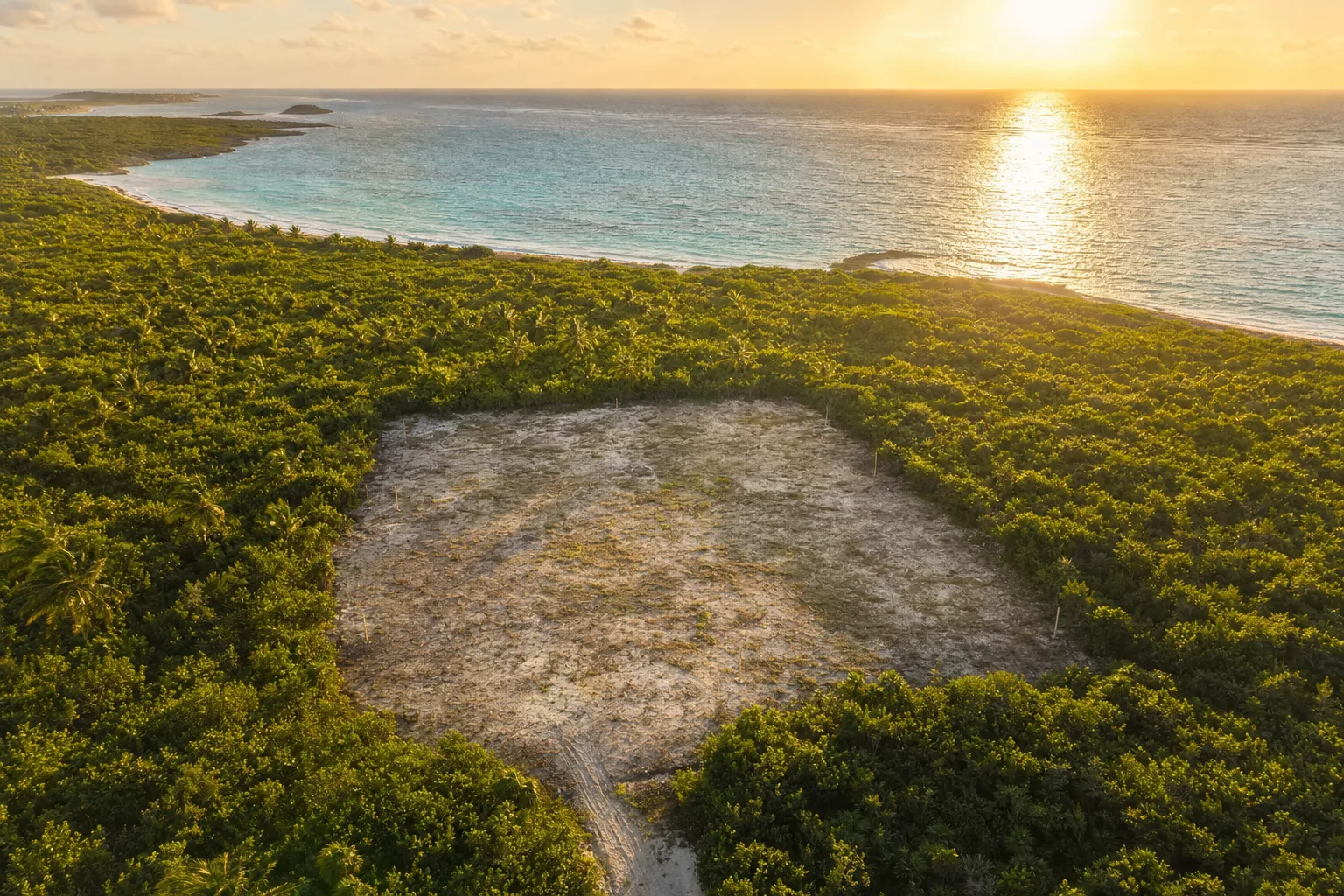 Vista aérea de un lote costero desmontado al atardecer, con las estacas de mensura en las esquinas, la selva alrededor y el mar turquesa detrás