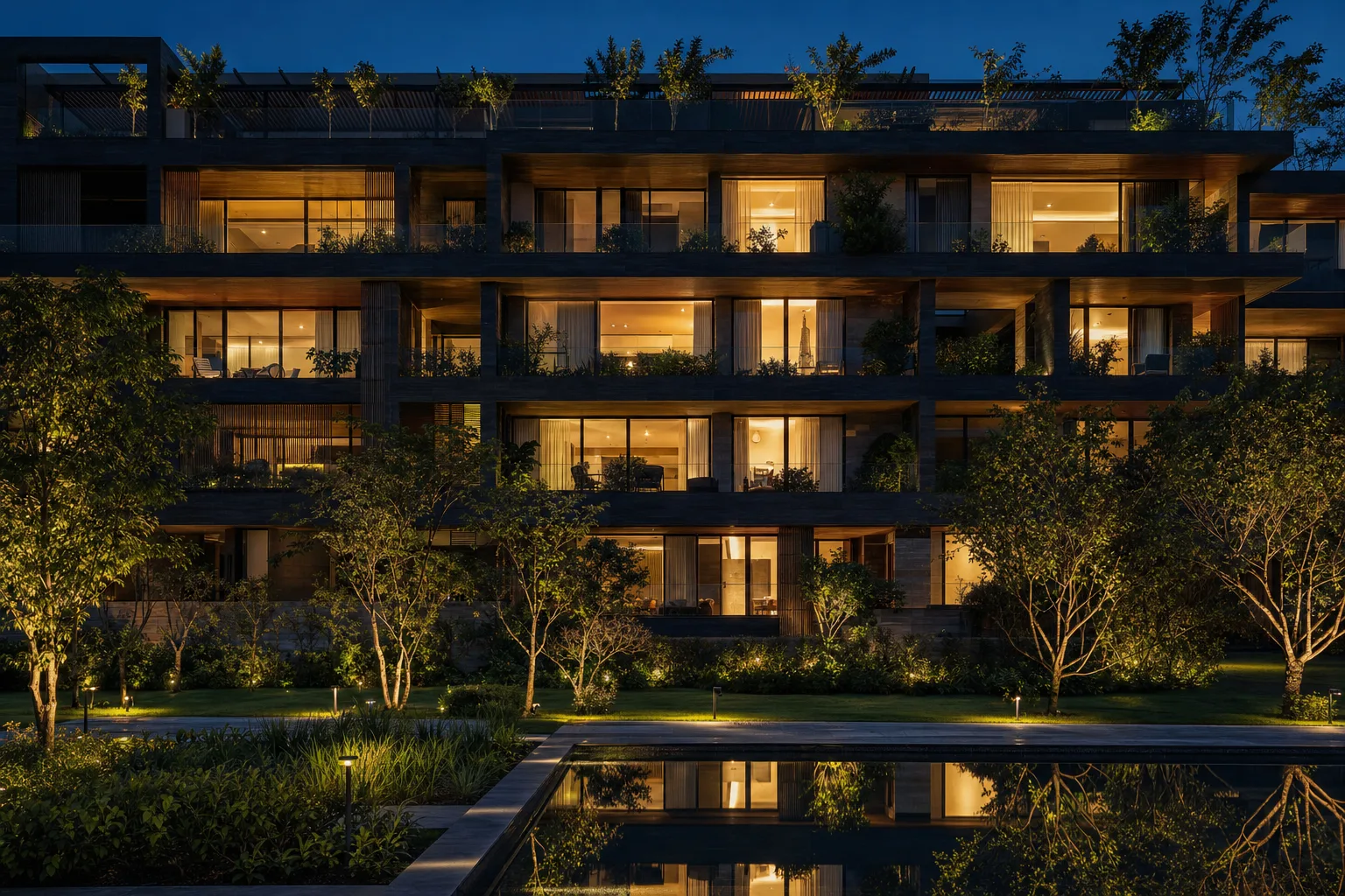 Edificio residencial de noche visto desde el jardín, con la mayoría de los departamentos encendidos y el espejo de agua reflejándolos